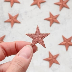 May include: A hand holding a rose gold star-shaped patch. The patch is made of fabric and has a quilted texture.
