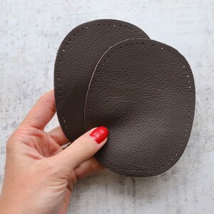May include: Two brown leather patches with stitched edges. The patches are oval shaped and have a textured surface.