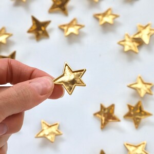 Puede incluir: Una mano sostiene un adorno dorado en forma de estrella. La estrella es texturizada y tiene un aspecto ligeramente martillado. Varias otras estrellas dor&eacute;es est&aacute;n esparcidas sobre una superficie blanca.