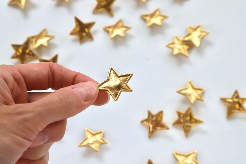 Puede incluir: Una mano sostiene un adorno dorado en forma de estrella. La estrella es texturizada y tiene un aspecto ligeramente martillado. Varias otras estrellas dor&eacute;es est&aacute;n esparcidas sobre una superficie blanca.
