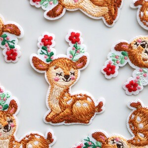 May include: A set of eight embroidered iron-on patches featuring cute deer with floral accents. The deer are brown with white spots and have closed eyes. The flowers are red, green, and white.