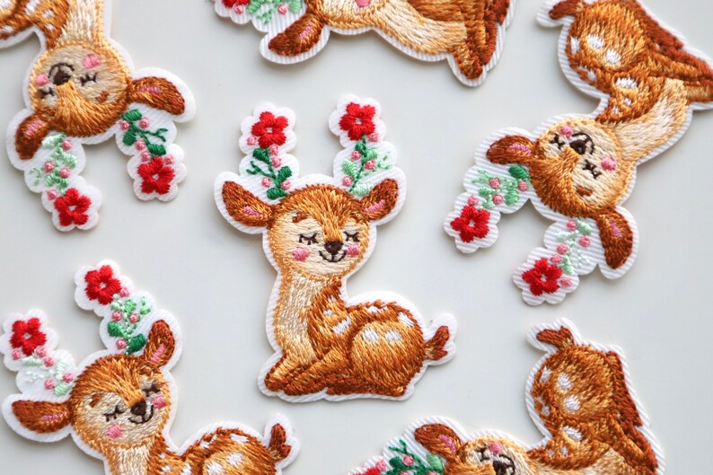 May include: A set of eight embroidered iron-on patches featuring cute deer with floral accents. The deer are brown with white spots and have closed eyes. The flowers are red, green, and white.