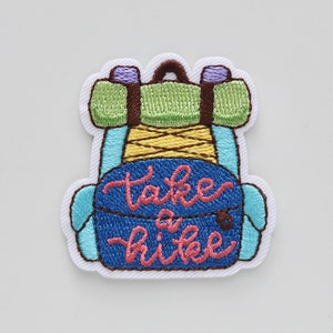 May include: Embroidered patch with a blue backpack and the text "take a hike" in pink. The backpack has a green sleeping bag and yellow straps.