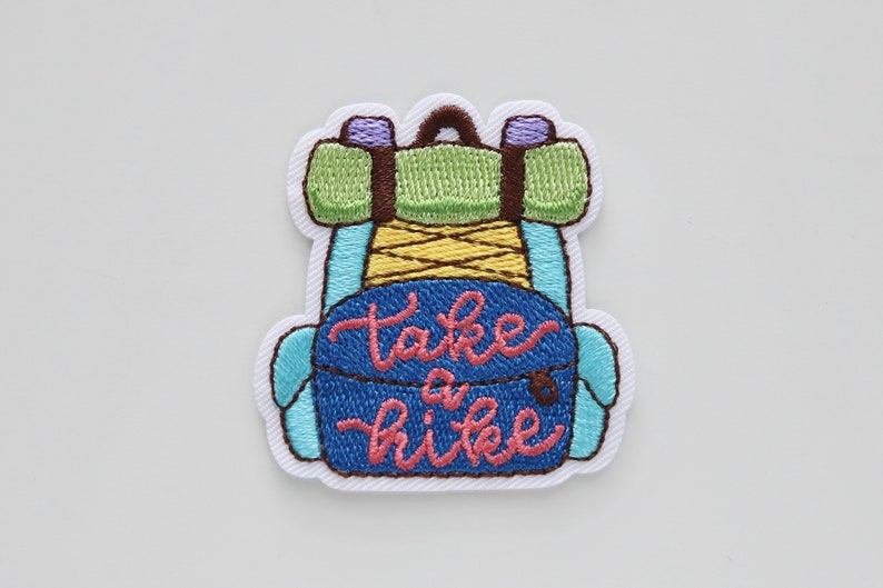 May include: Embroidered patch with a blue backpack and the text "take a hike" in pink. The backpack has a green sleeping bag and yellow straps.