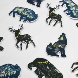 May include: A set of ten iron-on patches featuring various animals and nature scenes. The patches are black, green, and blue with a detailed, intricate design. The animals include a wolf, a deer, a bear, and a buffalo.