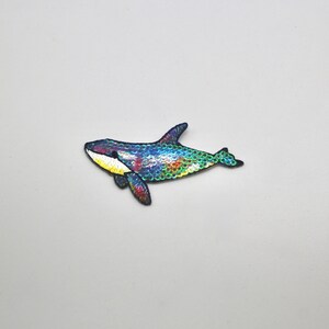May include: A colorful iridescent blue, green, and yellow embroidered patch of an orca whale. The patch is perfect for adding a whimsical touch to clothing, bags, or other accessories.