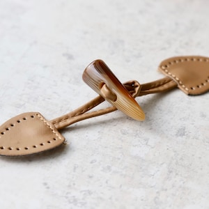 May include: A brown leather toggle closure with a brown horn toggle. The toggle is attached to two leather loops.