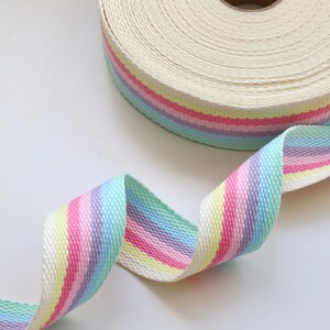May include: A white cotton webbing ribbon with pastel rainbow stripes. The ribbon is rolled up and a section is unfurled, showing the stripes.