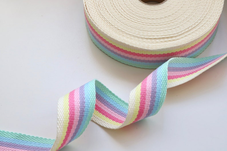 May include: A white cotton webbing ribbon with pastel rainbow stripes. The ribbon is rolled up and a section is unfurled, showing the stripes.