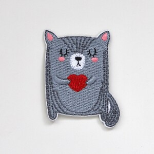 May include: Embroidered iron-on patch of a gray cat holding a red heart. The cat has pink cheeks and closed eyes.
