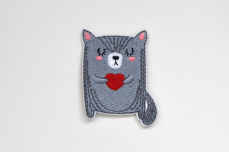May include: Embroidered iron-on patch of a gray cat holding a red heart. The cat has pink cheeks and closed eyes.