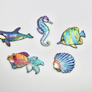 May include: Five colourful, sparkly, iron-on patches featuring a seahorse, a fish, a seashell, a whale, and a goldfish. These patches are perfect for adding a touch of whimsy to clothing, bags, or other fabric items.