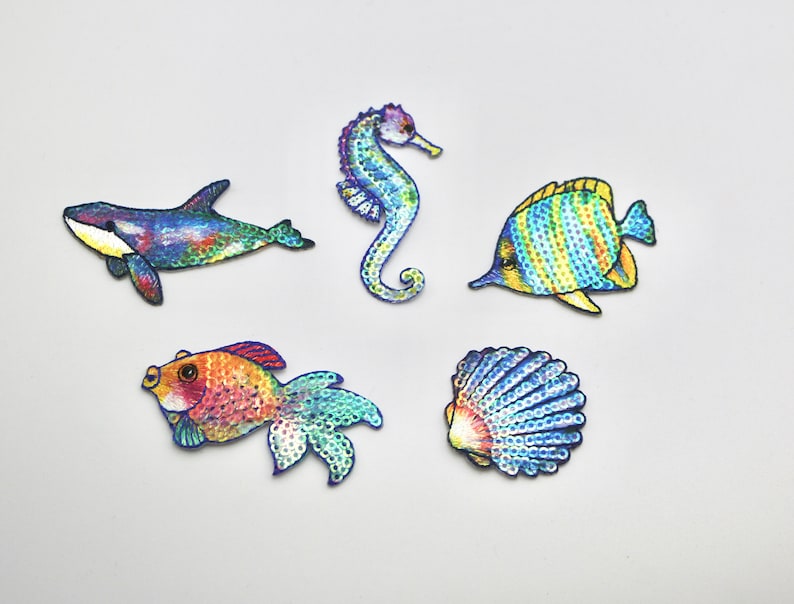 May include: Five colorful, sparkly, iron-on patches featuring a seahorse, a fish, a seashell, a whale, and a goldfish. These patches are perfect for adding a touch of whimsy to clothing, bags, or other fabric items.
