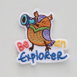 May include: Embroidered iron-on patch featuring a cartoon owl wearing a green and purple vest and holding a blue telescope. The owl is standing on a white background with the text "Be an Explorer" in blue, yellow, and pink.