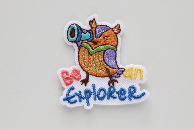 May include: Embroidered iron-on patch featuring a cartoon owl wearing a green and purple vest and holding a blue telescope. The owl is standing on a white background with the text "Be an Explorer" in blue, yellow, and pink.