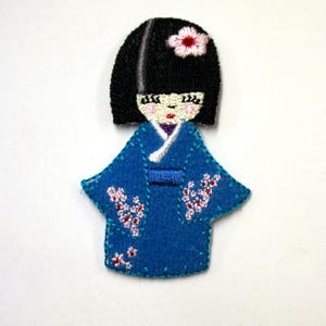 May include: Embroidered felt patch of a Japanese doll wearing a blue kimono with pink flowers. The doll has black hair and a pink flower on her head.