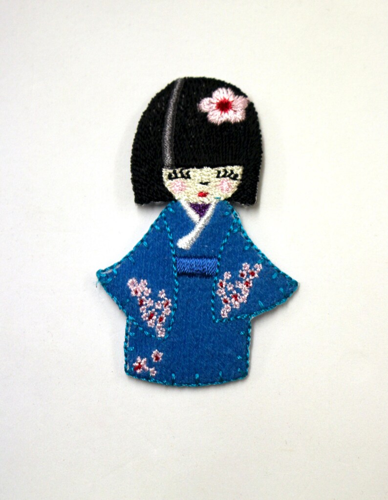 May include: Embroidered felt patch of a Japanese doll wearing a blue kimono with pink flowers. The doll has black hair and a pink flower on her head.