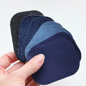 May include: A hand displays a selection of denim fabric swatches in shades of blue and black. The oval-shaped swatches are arranged to show the different colours and textures of the denim material.