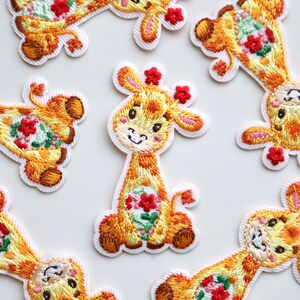 May include: Nine embroidered iron-on patches of yellow giraffes with red flowers. The giraffes have pink ears and are smiling.
