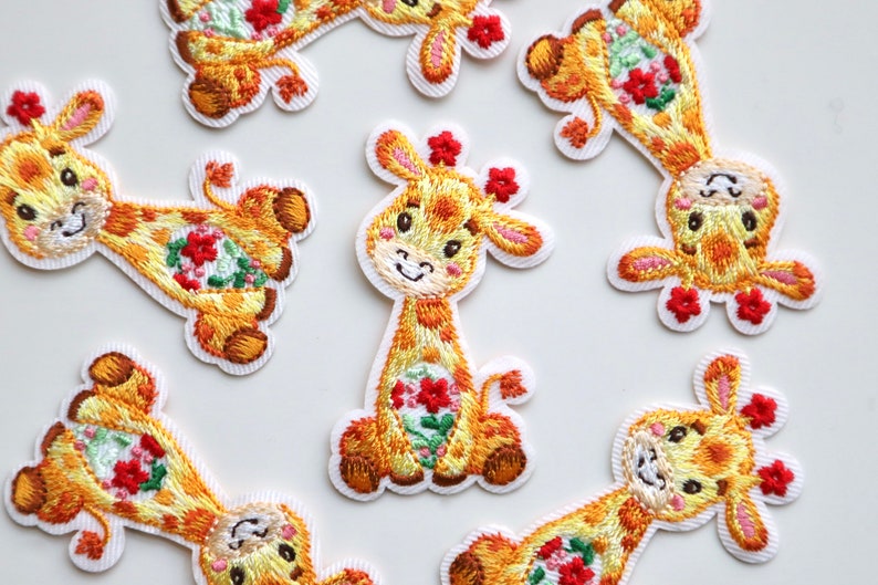 May include: Nine embroidered iron-on patches of yellow giraffes with red flowers. The giraffes have pink ears and are smiling.