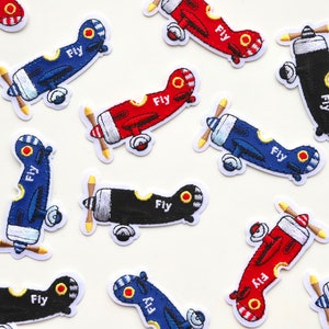 May include: Assortment of embroidered iron-on patches featuring aeroplanes in red, blue, and black. Each patch has the word "Fly" written on it.