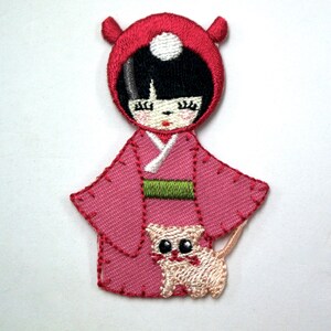 May include: Embroidered patch of a girl wearing a red hooded dress and holding a small cat. The girl has black hair and closed eyes. The cat is white with black eyes.