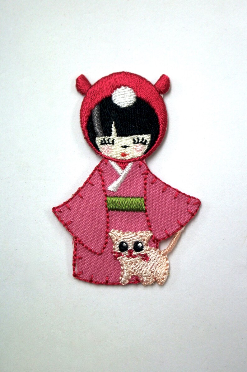 May include: Embroidered patch of a girl wearing a red hooded dress and holding a small cat. The girl has black hair and closed eyes. The cat is white with black eyes.