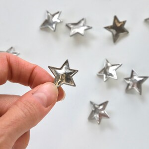 Puede incluir: Una colecci&oacute;n de nueve adornos met&aacute;licos en forma de estrella plateada sobre un fondo blanco. Una estrella est&aacute; siendo sostenida por una mano.
