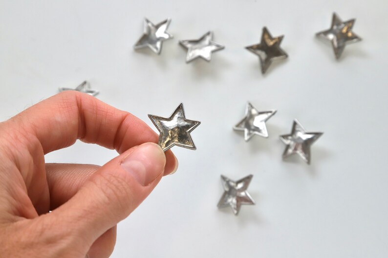 Puede incluir: Una colecci&oacute;n de nueve adornos met&aacute;licos en forma de estrella plateada sobre un fondo blanco. Una estrella est&aacute; siendo sostenida por una mano.