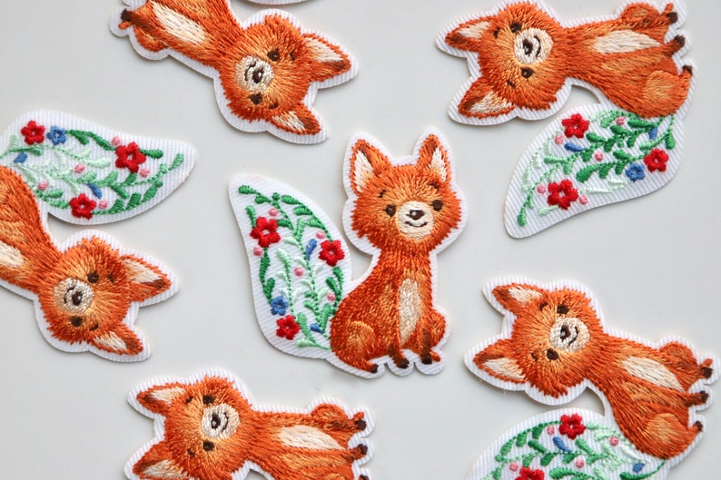 May include: A set of nine embroidered iron-on patches featuring foxes with floral details. The foxes are orange and brown with white accents. The flowers are red, blue, and green.