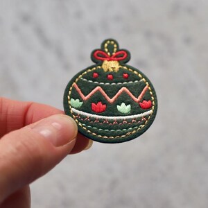 Peut inclure: Une d&eacute;coration de No&euml;l en feutre vert avec des broderies rouges et blanches. La d&eacute;coration est en forme de boule et pr&eacute;sente un motif en zigzag.