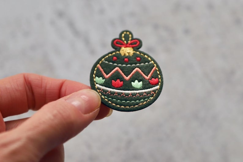 Peut inclure: Une d&eacute;coration de No&euml;l en feutre vert avec des broderies rouges et blanches. La d&eacute;coration est en forme de boule et pr&eacute;sente un motif en zigzag.