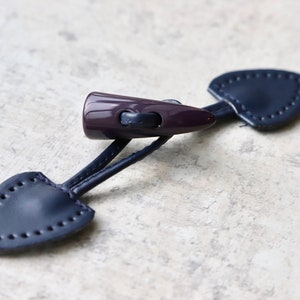 May include: A dark purple plastic toggle with two dark blue leather loops. The loops have decorative stitching around the edges.