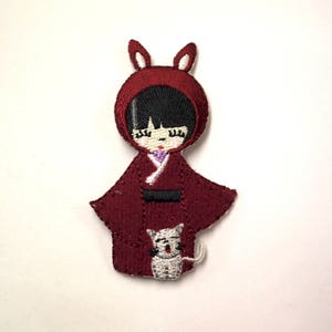 May include: Embroidered iron-on patch of a girl wearing a red hooded dress with bunny ears and holding a white cat.