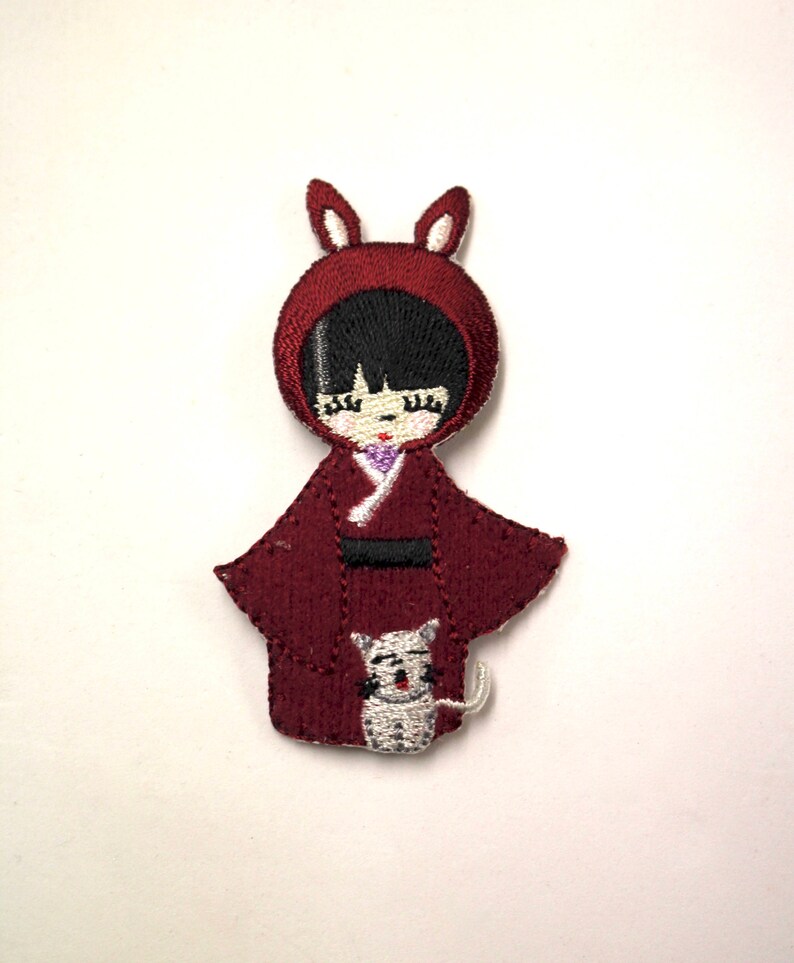 May include: Embroidered iron-on patch of a girl wearing a red hooded dress with bunny ears and holding a white cat.