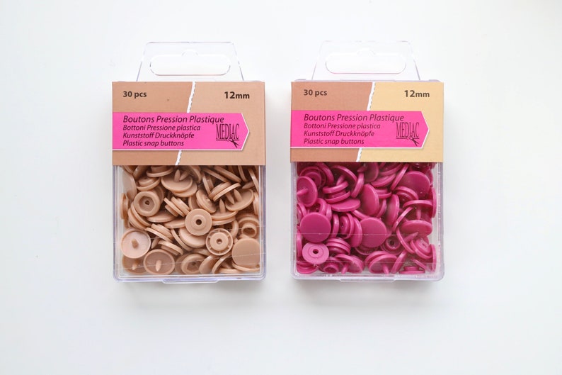 K&ouml;nnte beinhalten: Zwei Packungen mit je 30 Kunststoff-Druckkn&ouml;pfen, jeweils 12 mm gro&szlig;. Eine Packung enth&auml;lt beige Kn&ouml;pfe, die andere enth&auml;lt rosa Kn&ouml;pfe. Die Packungen sind mit "Boutons Pression Plastique" und "MEDIAC" beschriftet.