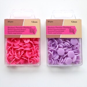 K&ouml;nnte beinhalten: Zwei Packungen mit je 30 Kunststoff-Druckkn&ouml;pfen, jeweils 12 mm gro&szlig;. Die Packung links enth&auml;lt rosa Kn&ouml;pfe, w&auml;hrend die Packung rechts lila Kn&ouml;pfe enth&auml;lt. Beide Packungen tragen den Text "Boutons Pression Plastique" und "MEDIAC".