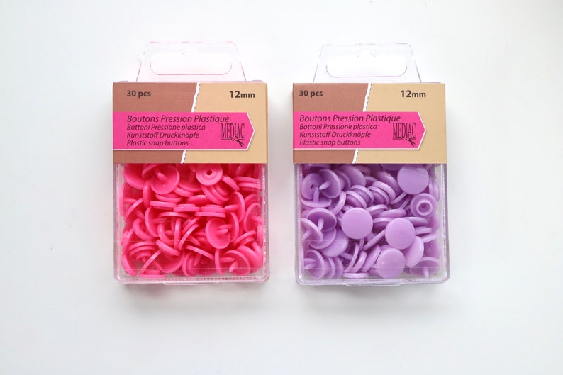 K&ouml;nnte beinhalten: Zwei Packungen mit je 30 Kunststoff-Druckkn&ouml;pfen, jeweils 12 mm gro&szlig;. Die Packung links enth&auml;lt rosa Kn&ouml;pfe, w&auml;hrend die Packung rechts lila Kn&ouml;pfe enth&auml;lt. Beide Packungen tragen den Text "Boutons Pression Plastique" und "MEDIAC".