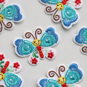 May include: Five embroidered iron-on patches featuring blue and turquoise butterflies with red, yellow, and green accents. The butterflies have a whimsical design with floral details.