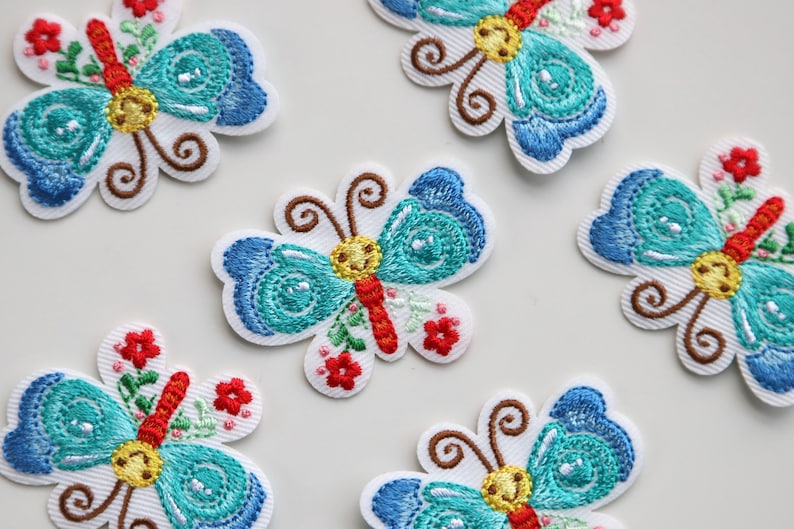 May include: Five embroidered iron-on patches featuring blue and turquoise butterflies with red, yellow, and green accents. The butterflies have a whimsical design with floral details.