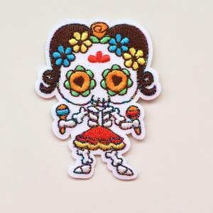 May include: Embroidered iron-on patch featuring a skeleton girl with brown hair, wearing a red and orange dress and holding maracas. She has a skull face with colourful flowers and a red dot on her forehead.