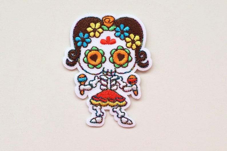 May include: Embroidered iron-on patch featuring a skeleton girl with brown hair, wearing a red and orange dress and holding maracas. She has a skull face with colourful flowers and a red dot on her forehead.