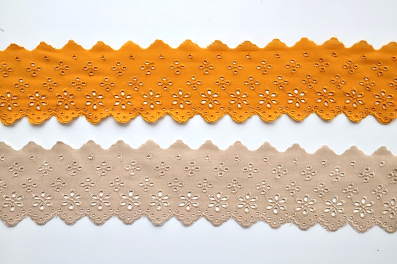 May include: Two pieces of scalloped cotton lace trim with a floral pattern. The top piece is mustard yellow and the bottom piece is beige.