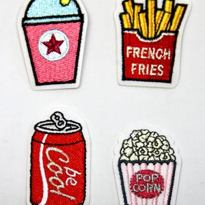 May include: Four embroidered iron-on patches featuring a red can of soda with the word "Coca-Cola" on it, a pink slushie with a straw and a red star, a red box of french fries with the words "French Fries" on it, and a white box of popcorn with the words "Pop Corn" on it.