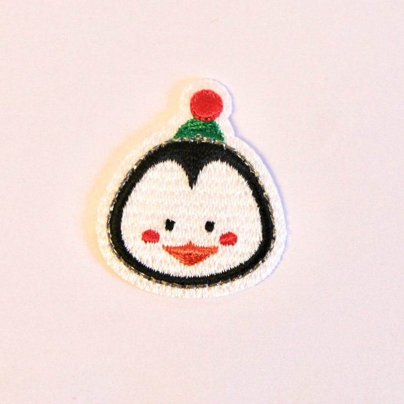 K&ouml;nnte beinhalten: Wei&szlig;er gestickter Pinguin-Patch mit roter und gr&uuml;ner M&uuml;tze. Der Pinguin hat schwarze Augen, einen schwarzen Schnabel und rote Wangen.