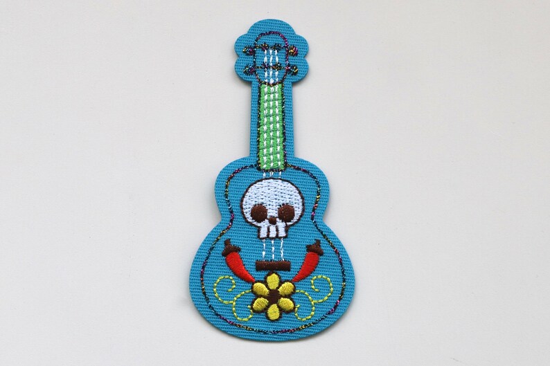 May include: A blue embroidered guitar with a skull and flower design. The guitar has a green neck and red chilli peppers on the body.