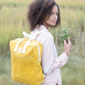 May include: A young person with curly hair is wearing a light pink shirt and a yellow canvas backpack. They are holding a bunch of white wildflowers.