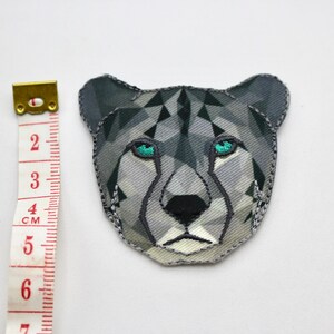 May include: A gray and white geometric cheetah head patch with teal eyes. The patch is approximately 6 cm wide.