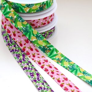 Puede incluir: Tres rollos de cinta estampada. La primera cinta es verde con un estampado de frutas tropicales. La segunda cinta es rosa con lunares blancos y fresas rojas. La tercera cinta es morada con un estampado floral.