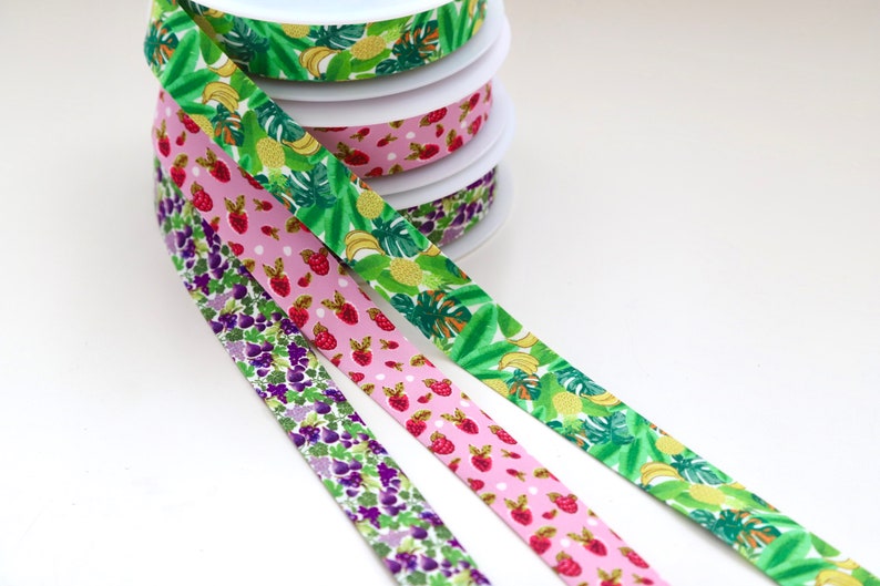 Puede incluir: Tres rollos de cinta estampada. La primera cinta es verde con un estampado de frutas tropicales. La segunda cinta es rosa con lunares blancos y fresas rojas. La tercera cinta es morada con un estampado floral.